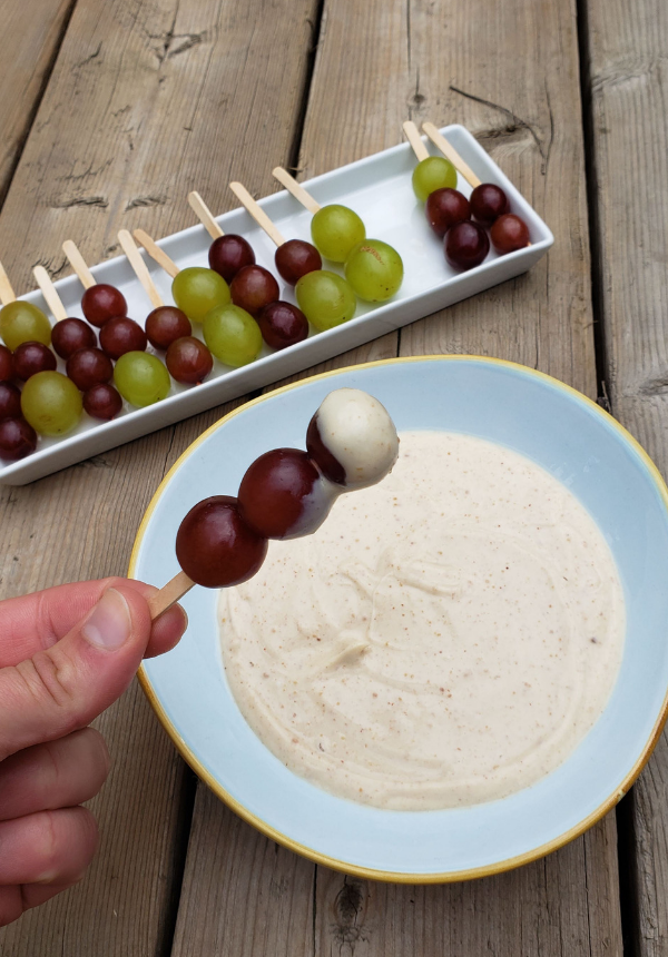 Grapes on popsicle sticks are on a tray waiting to be dipped into the bowl of peanut butter dip