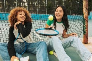 Two teens sitting on a tennis court playing with tennis ball and rackets